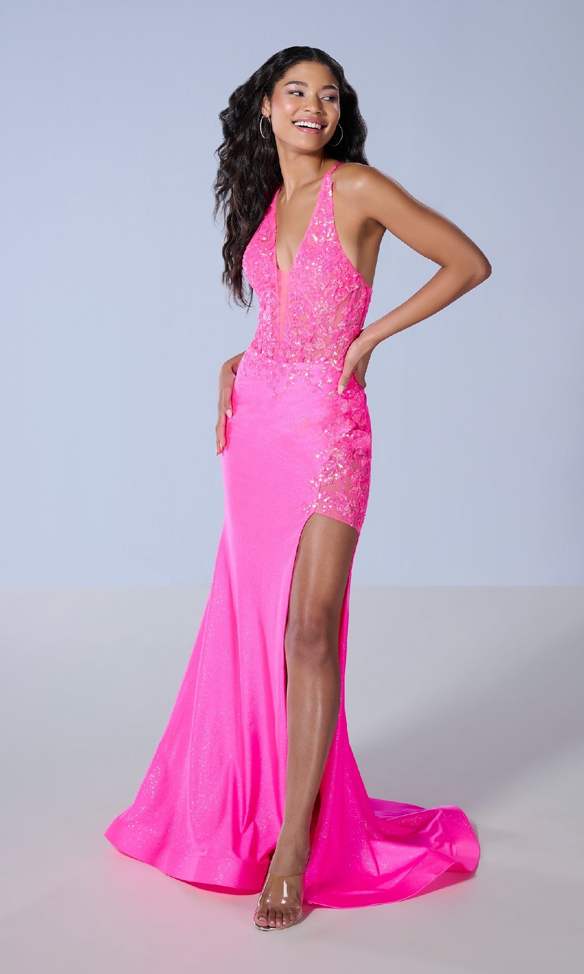 A woman stands against a plain light gray background wearing the Long Prom Dress: Tiffany 16246, featuring bright pink sequins, a corset bodice, and a thigh-high slit.
